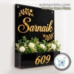 Black A4 acrylic house name plate with custom gold lettering "Sarnaik" and an integrated planter box holding green plants below house number 609.